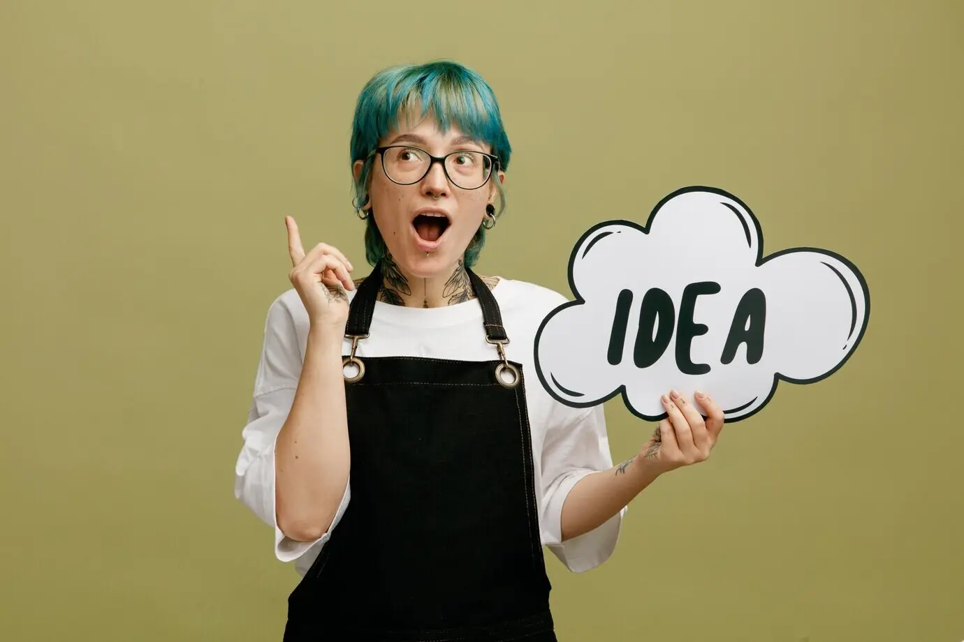 Inspirierte junge Barbierin mit Brille in Uniform, die eine Ideenblase zeigt, nach oben blickt und nach oben deutet, freigestellt vor olivgrünem Hintergrund.